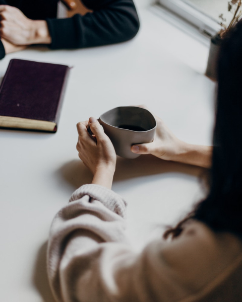 Cette photo représente un rendez-vous en présentiel entre deux personnes autour d'une table. Au premier plan, nous devinons une femme aux longs cheveux bruns sur le côté de l'image, tenant un mug entre ces mains et discutant avec une autre personne en face d'elle. Au second plan, en plus du mug, un ouvrage papier fermé est positionné devant la personne dont les mains croisées apparaissent au troisième plan.