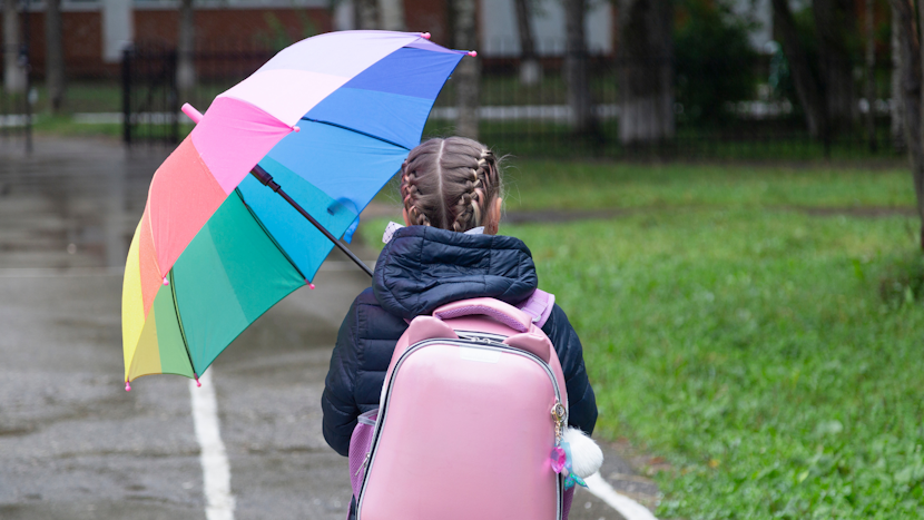 Elevă în ploaie, cu un ghiozdan în spate și o umbrelă în mână