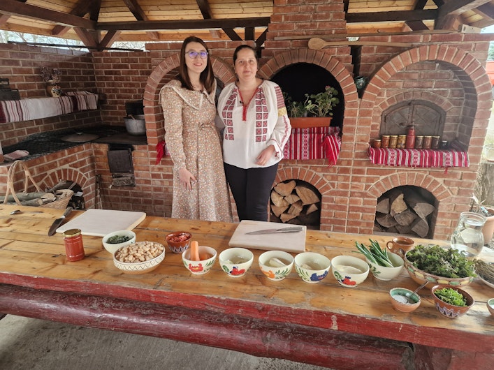 Două femei într-o bucătărie rustică din cărămidă, îmbrăcate în haine tradiționale