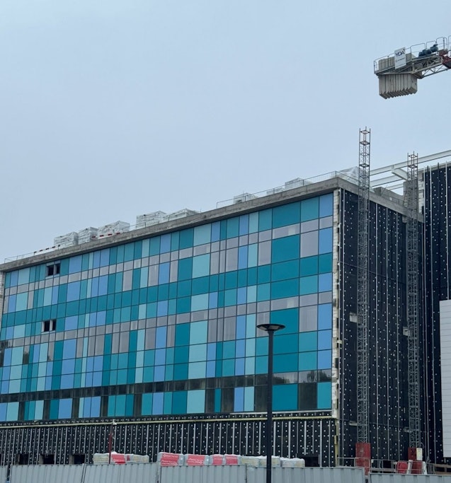 Clădire modernă aflată în construcție, cu fațadă din panouri de sticlă albastre și turcoaz