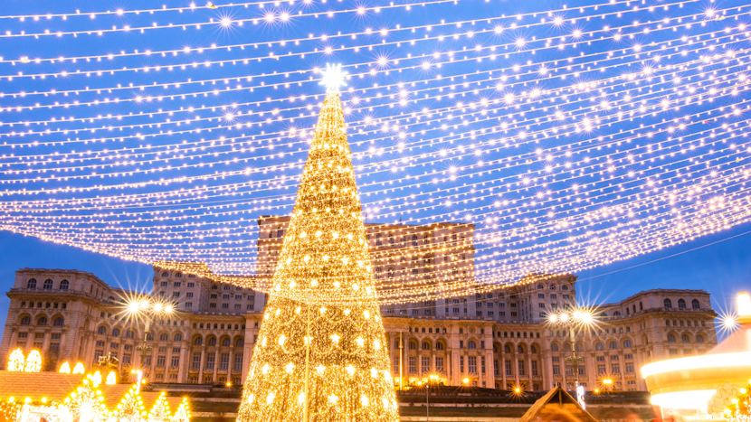 Un brad uriaș de Crăciun decorat cu mii de lumini aurii în centrul unui târg festiv, cu ghirlande luminoase întinse deasupra și o clădire mare în fundal, sub un cer albastru de seară.