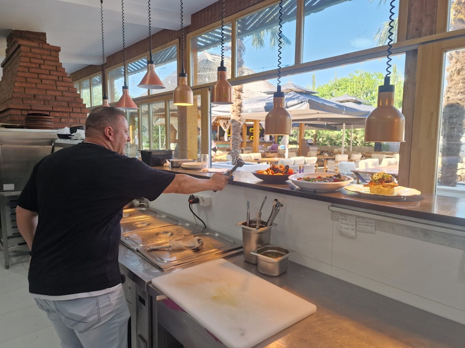 Bucătar într-o bucătărie deschisă, pregătind preparate pentru servire într-un restaurant cu terasă însorită