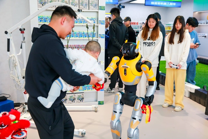 Robot umanoid se duce la un bebelus tinut in brate de tatal sau, alte persoane pe langa