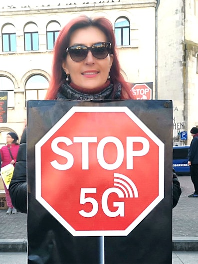Angela Negrotă, cu ochelari de soare, ținând în mână o pancartă cu un octogon roșu pe care este scris mesajul STOP 5G