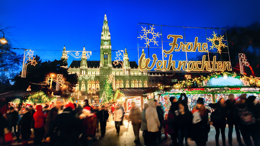 Târgul de Crăciun din fața primăriei vieneze prinde viață odată cu lăsarea serii: lumini calde, standuri colorate și mulțimea care se plimbă printre decorațiuni, sub urarea strălucitoare Frohe Weihnachten.