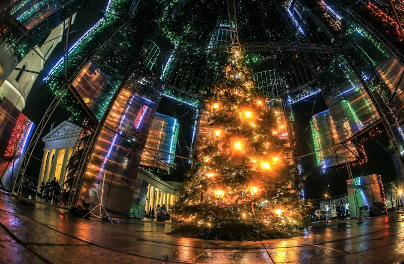 Un brad de Crăciun luminat stă în mijlocul unei instalații cu panouri reflectorizante și lumini colorate. Fotografia este făcută seara, iar reflexiile intensifică atmosfera festivă.