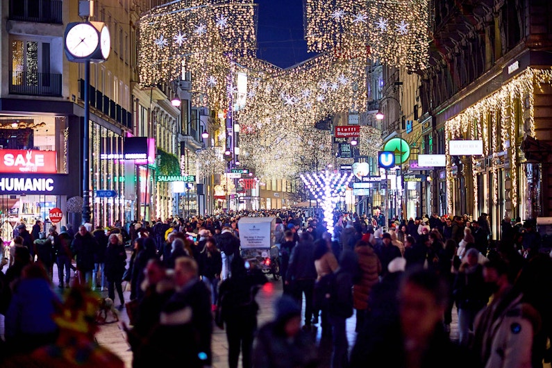 O stradă aglomerată, decorată cu lumini de Crăciun și ornamente strălucitoare suspendate deasupra trecătorilor. O mulțime de oameni se plimbă printre magazine luminate și vitrine festive, creând o atmosferă animată de sărbătoare.