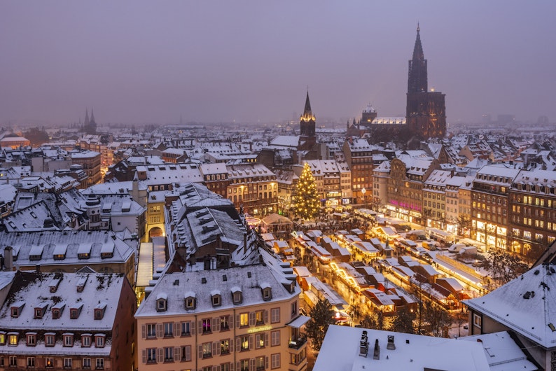 Un oraș acoperit de zăpadă văzut de sus, cu clădiri istorice și acoperișuri albe întinse până în depărtare. În centru se vede un târg de Crăciun luminat și un brad uriaș decorat, creând un contrast cald în peisajul de iarnă.
