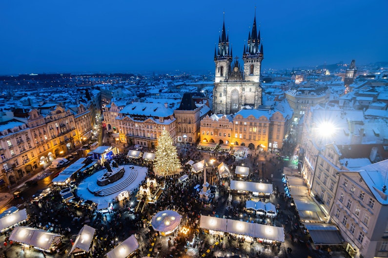 O piață mare acoperită de zăpadă, plină de căsuțe luminate și un brad de Crăciun în centru. În fundal se văd clădiri istorice și o catedrală impunătoare, toate sub cerul albastru al serii.