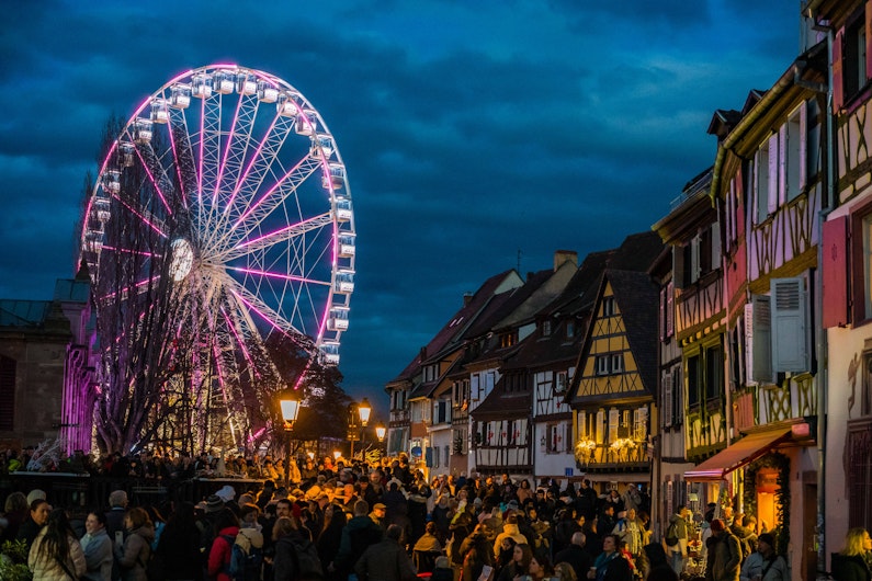 Un târg de Crăciun plin de vizitatori, cu căsuțe luminate și decorațiuni aurii atârnate deasupra. Clădiri elegante și un brad împodobit se văd în fundal, creând o atmosferă festivă de seară.