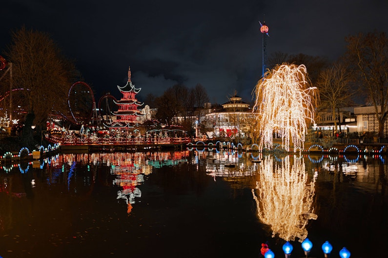 O clădire în stil pagodă și un copac acoperit cu lumini aurii se reflectă în apa unui lac, într-un parc decorat pentru sărbători. Iluminările colorate creează o atmosferă festivă pe fundalul cerului întunecat.