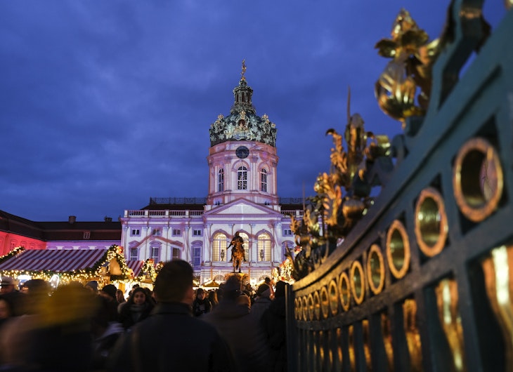Un palat baroc luminat în culori pastel stă în fundalul unui târg de Crăciun aglomerat. În prim-plan se vede poarta decorată cu detalii aurii, iar mulțimea se plimbă printre căsuțele luminate.