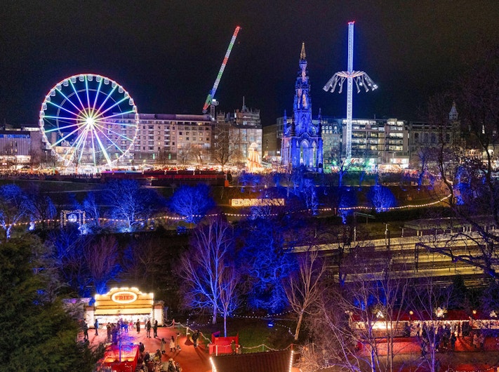 O roată panoramică colorată și mai multe atracții de parc se ridică deasupra unui târg de Crăciun iluminat. Copacii și clădirile din jur sunt scăldați în lumini albastre și aurii, creând un peisaj animat de seară.