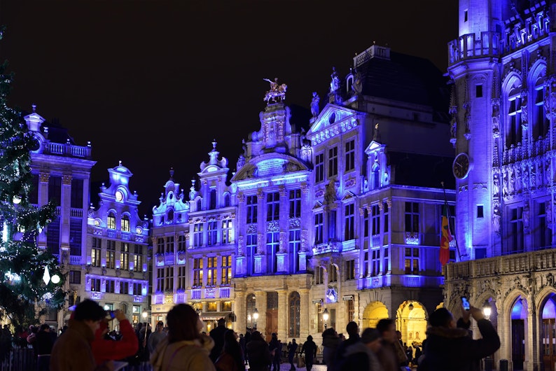 Fațade baroce iluminate în nuanțe de albastru domină o piață aglomerată seara. Oamenii se plimbă și fac fotografii, iar în stânga se vede un brad împodobit, adăugând un aer de sărbătoare.