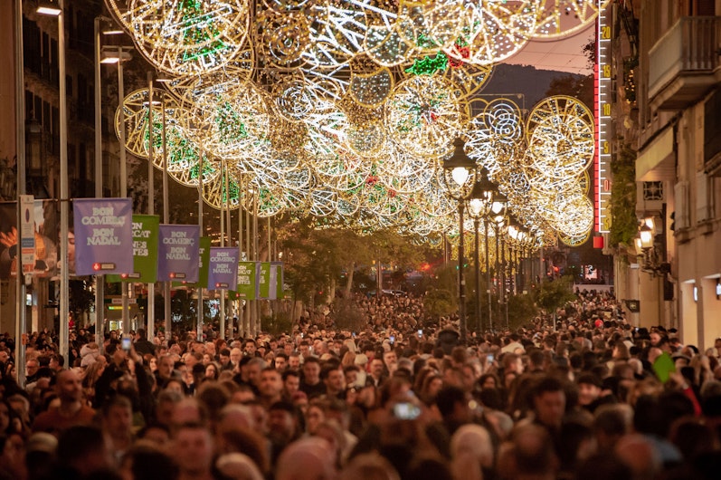 O mulțime densă umple o stradă decorată cu instalații luminoase elaborate în forme circulare, strălucind în alb, verde și auriu. Bannere festive și felinare se aliniază de-a lungul bulevardului, creând o atmosferă intensă de sărbătoare.