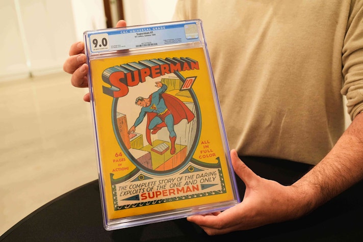 Un bărbat ține în mâini un exemplar rar și sigilat al revistei «Superman No. 1» (CGC 9.0), într-o carcasă transparentă de protecție, ilustrând coperta clasică cu Superman sărind peste clădiri.