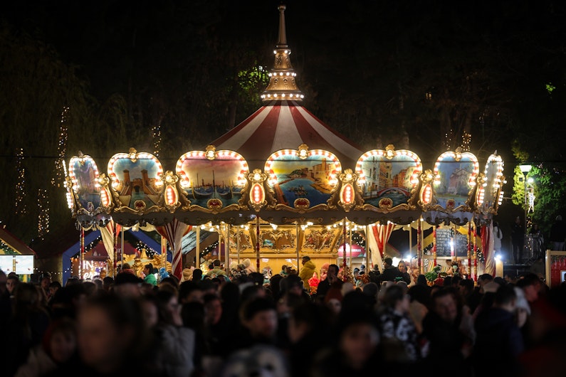 Carusel luminat într-un târg de Crăciun aglomerat, fotografiat noaptea, cu mulțimi de vizitatori și decoruri festive.