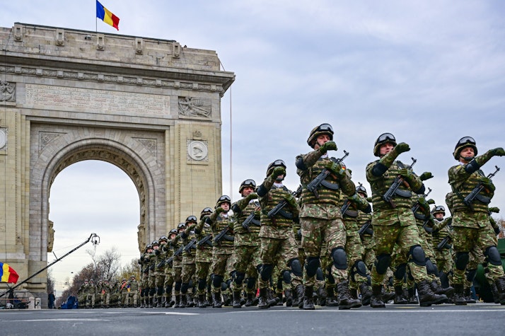Militari români în uniformă defilează în pas de paradă în fața Arcului de Triumf din București, cu drapelul României arborat deasupra monumentului.