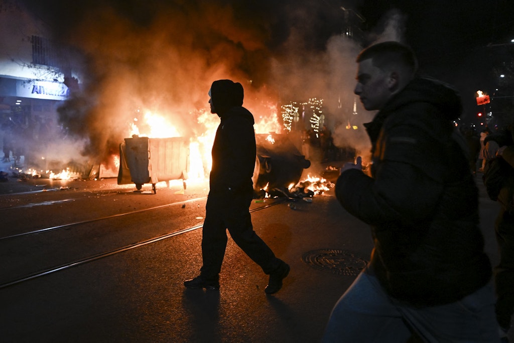 Un bărbat îmbrăcat în negru trece prin fața unor containere de gunoi în flăcări pe o stradă din Sofia, Bulgaria, în timpul unui protest care a degenerat în incendii și haos nocturn.
