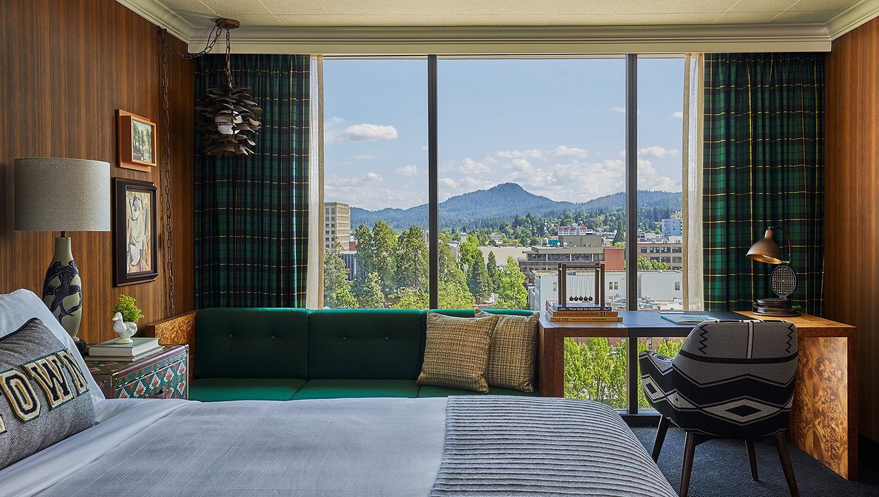 Side shot looking over a large hotel bed out to floor-to-ceiling windows with a beautiful day view of the city