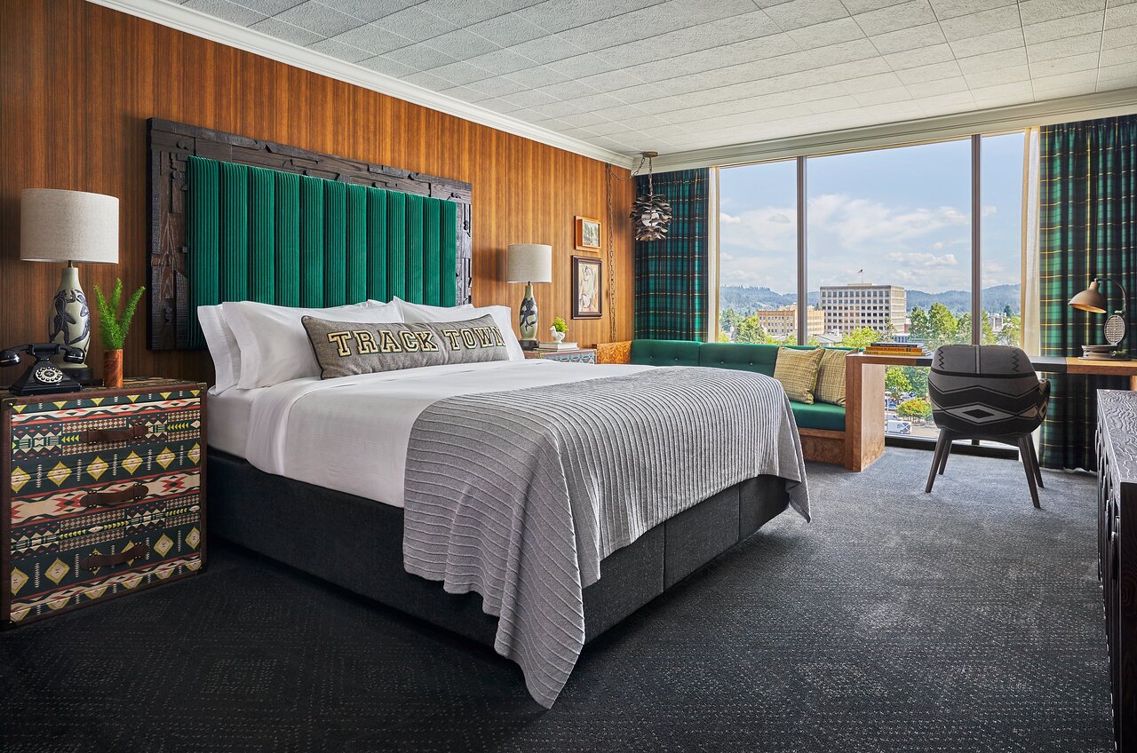 Off-to-the-side shot of large, comfortable-looking hotel bed with beautiful view of the city through the window in the background.