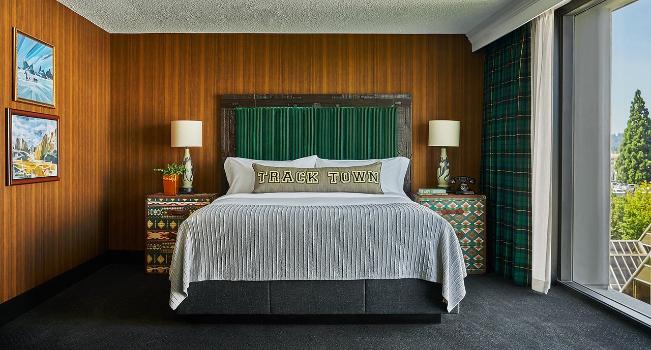 Large bed with green headboard in the focal point with wooden walls behind and large floor to ceiling windows to the right