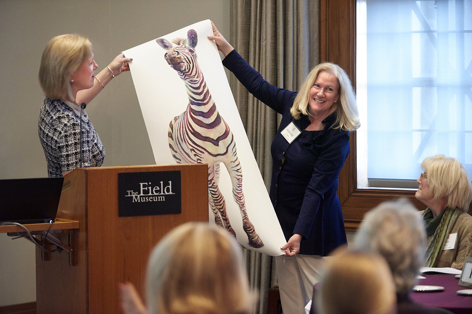 Two women hold up a painting of a zebra by Field Museum artist-in-residence Peggy McNamara, while standing behind a podium with the Museum’s logo on it. Other Women’s Board members, seated at tables, look on.