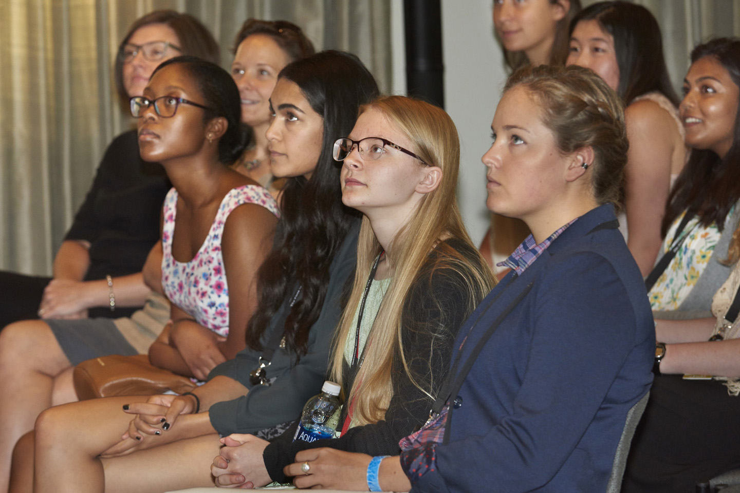 A row of four girls sit in a row and listen to a lecture. The speaker is not pictured.