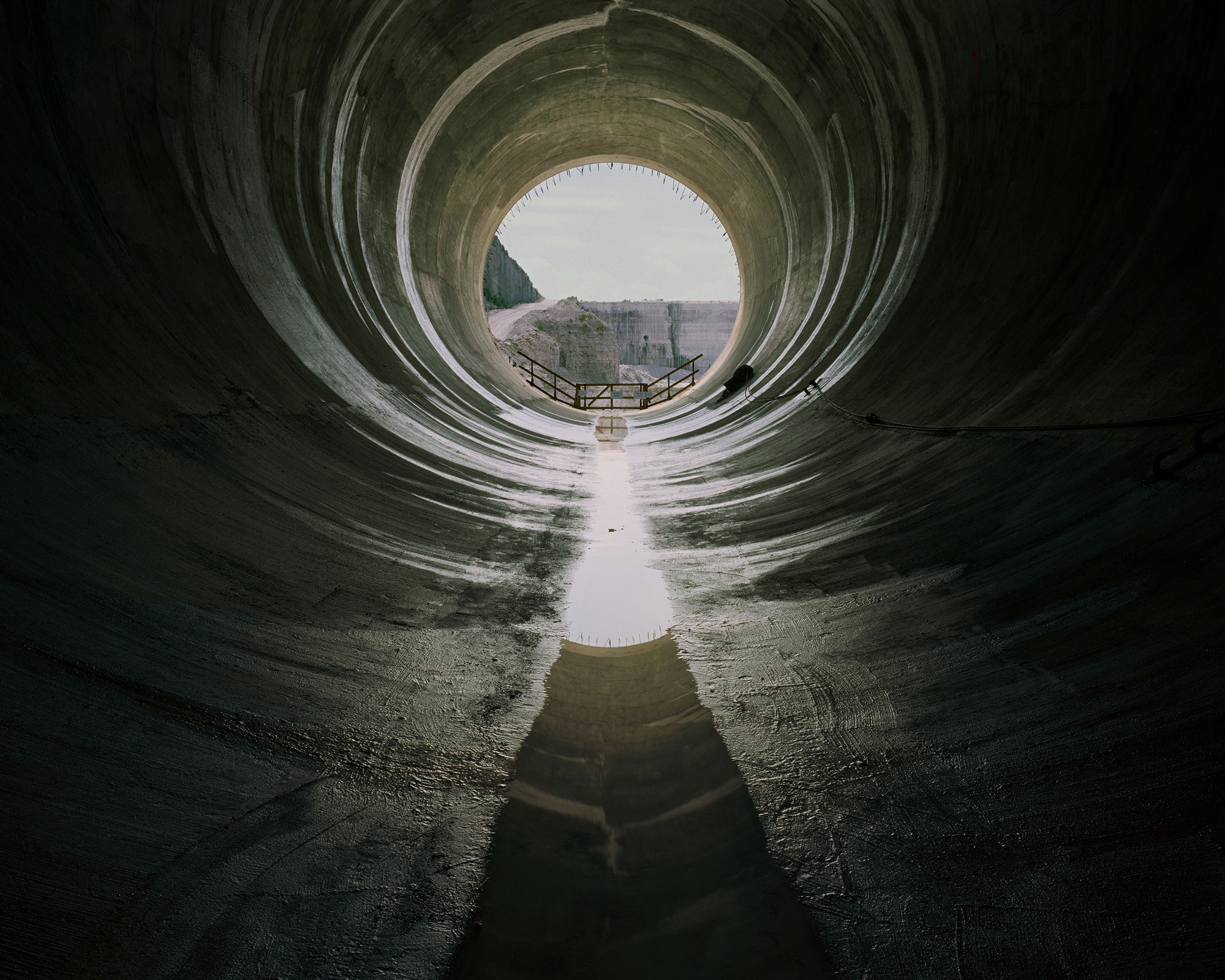 View from inside a large tunnel, looking out its circular opening in the distance. A shallow puddle runs the length of the tunnel.