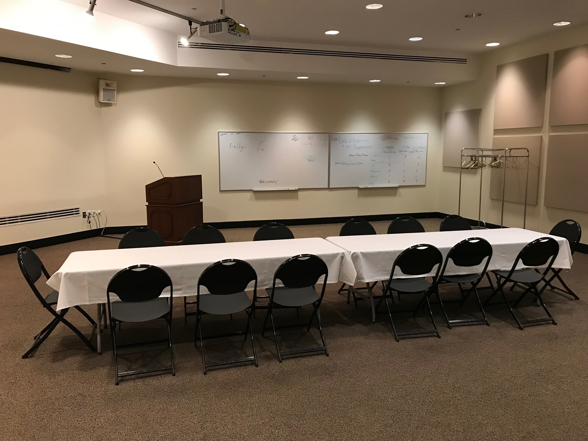 A long tables sits in the middle of Lecture Hall II. The table is set for 14 guests and topped with a white tablecloth. A podium, two whiteboards, and empty coat rack are visible in the back. A projector hangs overhead.