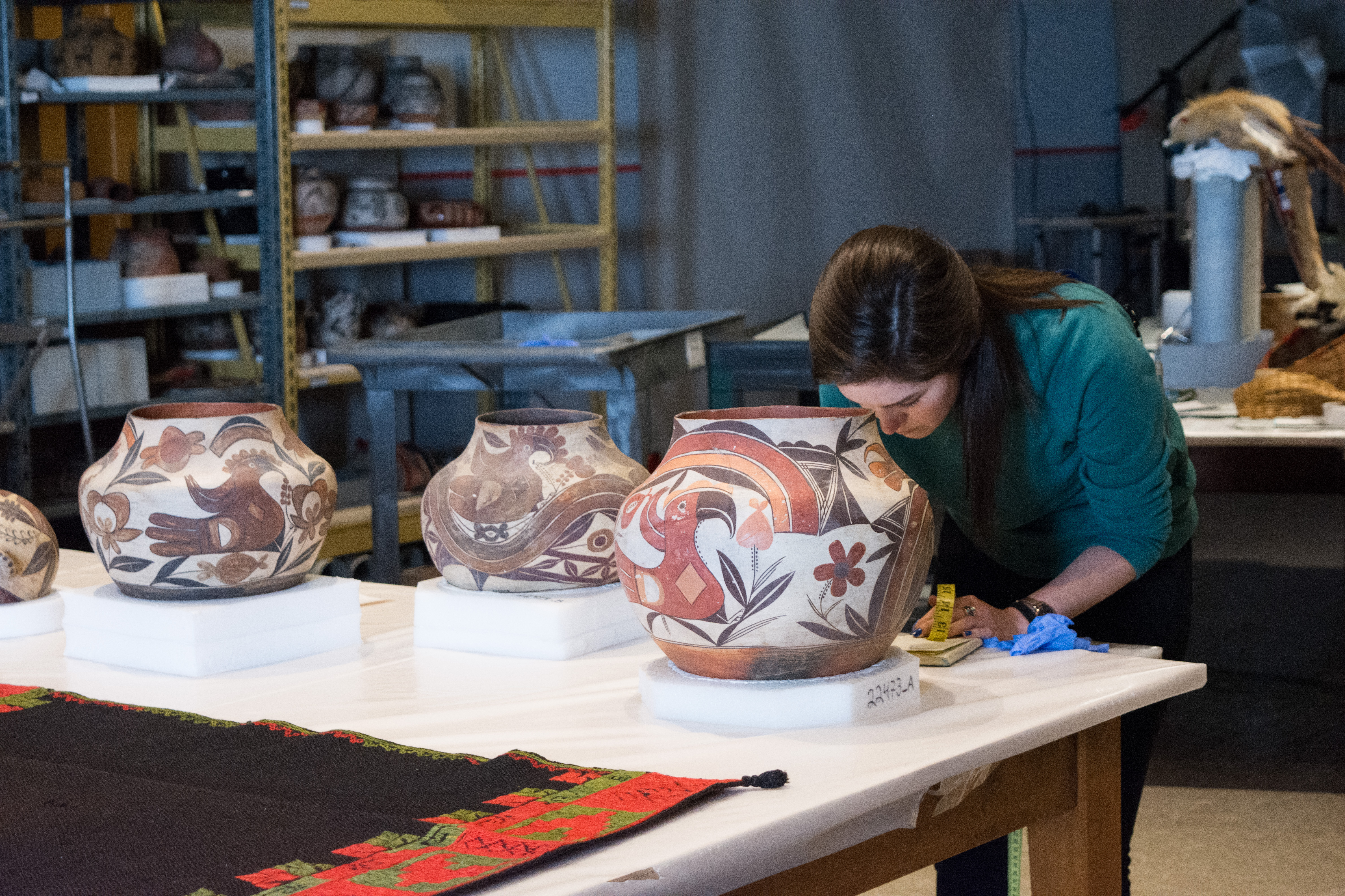 A woman stands behind a table with three large, painted pots. She holds a measuring tape and leans over the table to write something down.