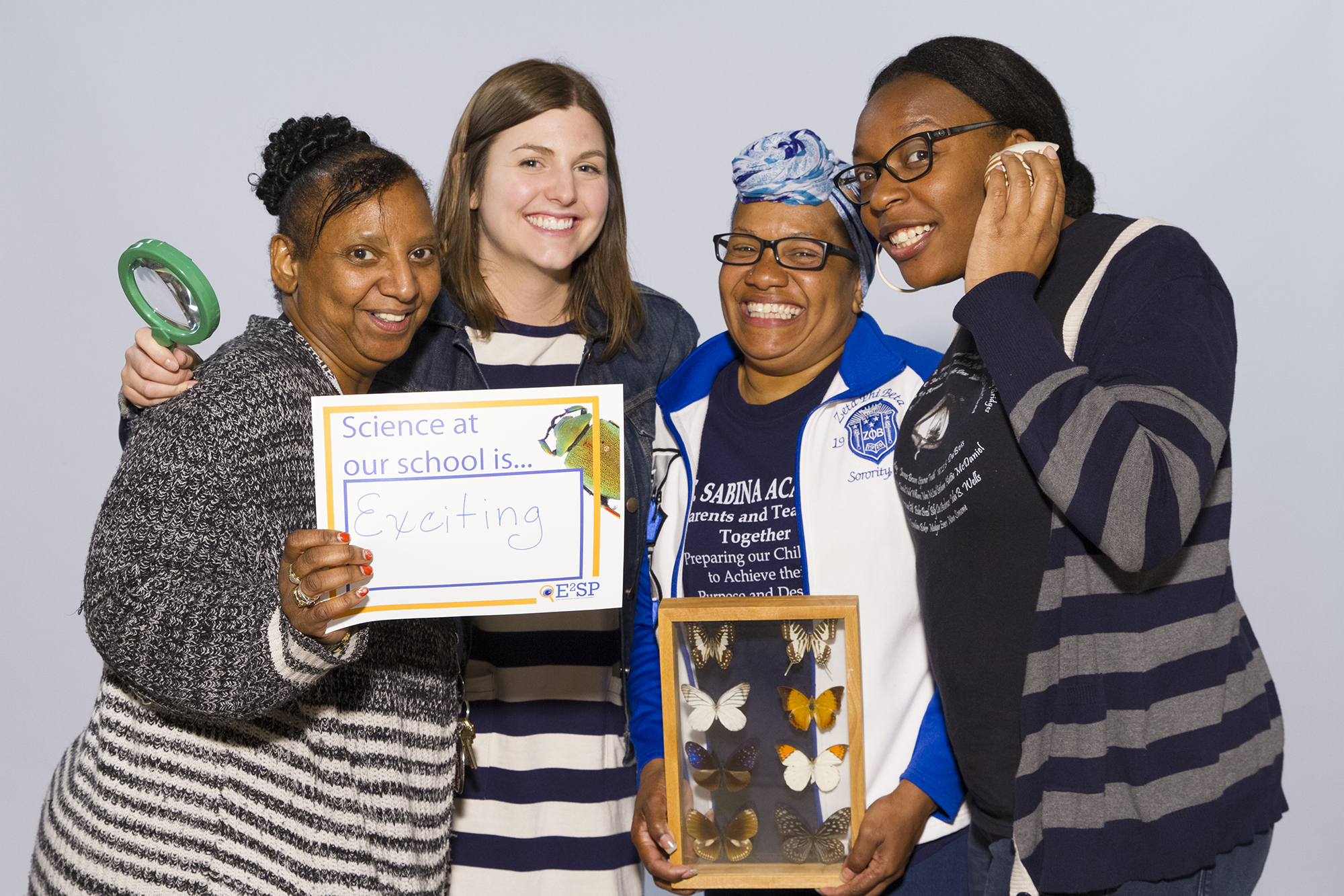 Four educators pose for a photo. One holds a sign that reads Science at our school is exciting. Two others hold a magnifying glass and a glass case of butterflies. The other smiles while holding her hand lightly up to her face.
