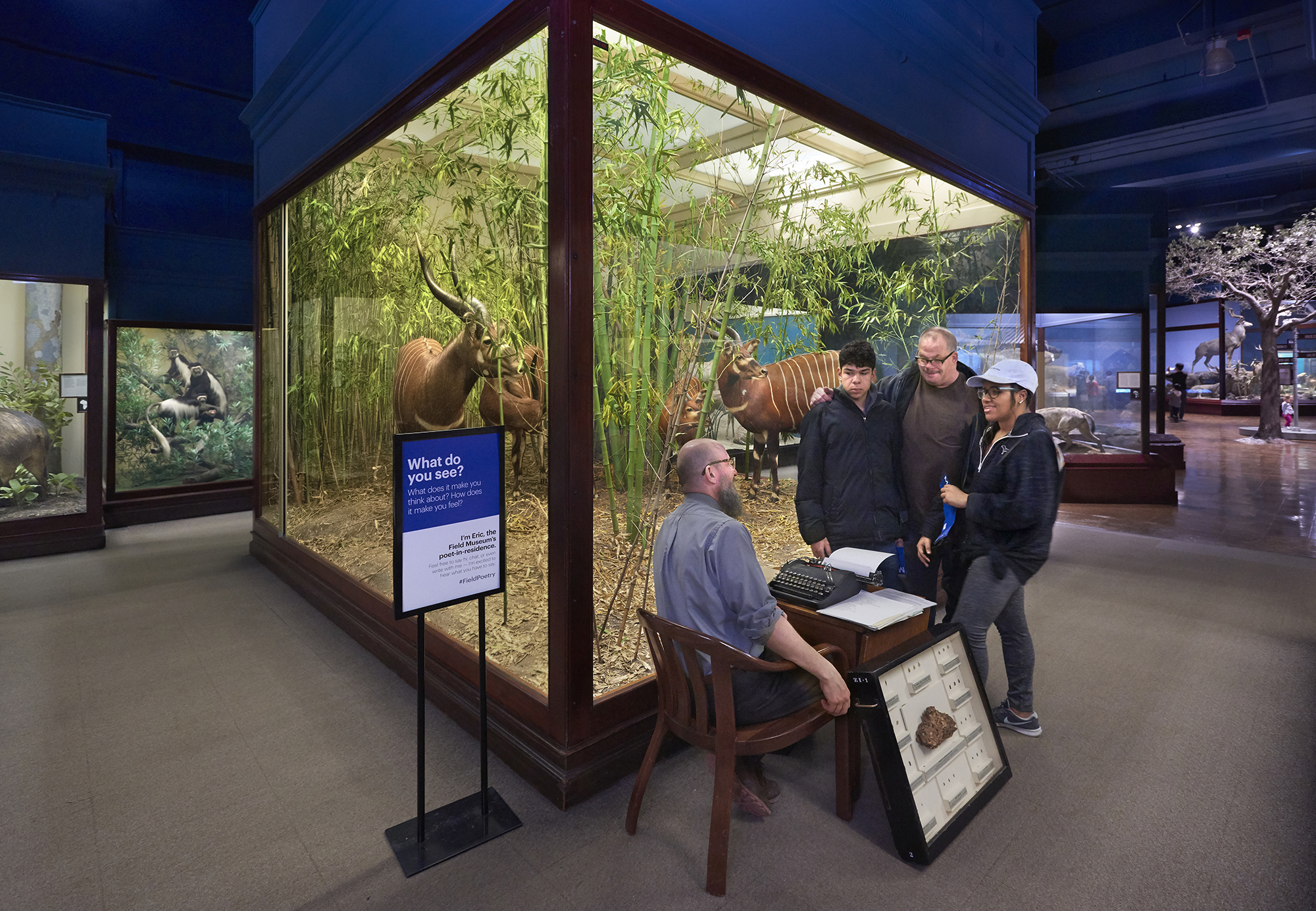 A group gathers at the corner of a diorama case. One man sits a desk with a typerwriter. He looks up to a family of three standing over him. The man in the middle stands with arms around the other two individuals. Both are smiling down at the man seated at the desk. A black box with a glass front holds a rock specimen and is propped up against the leg of the desk.