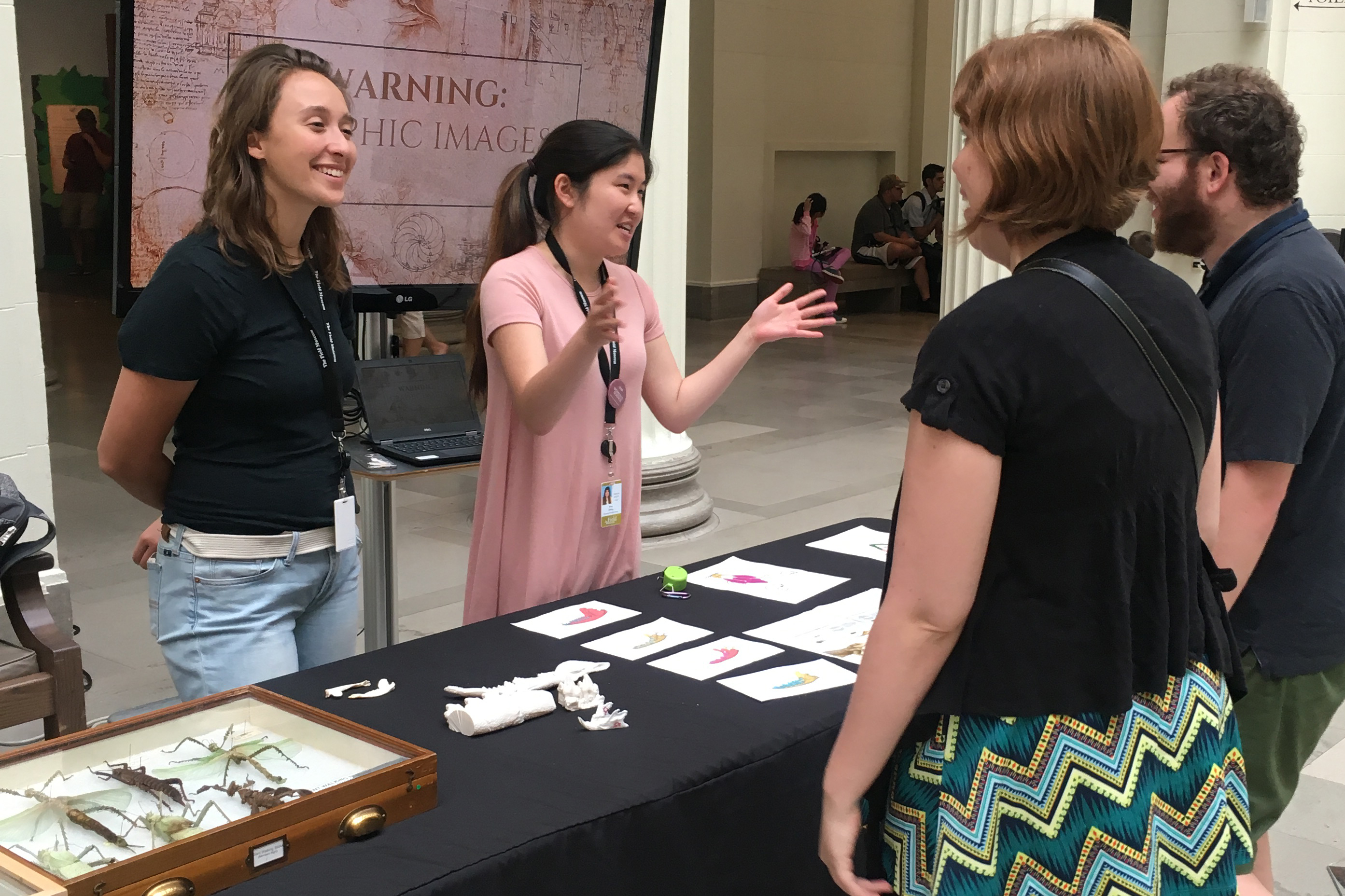 Two interns stand behind a table displaying objects from the Field Museum’s collection. One speaks with the two people in front of her, gesturing as if she’s showing the size of something.