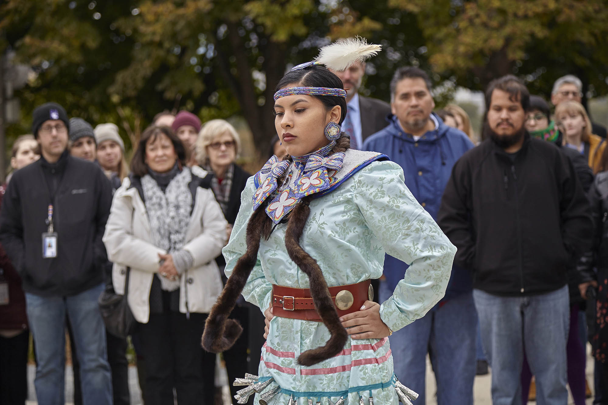A woman, Maritza Garcia, wearing a light blue dress and a headdress with a feather, performs a dance in front of onlookers at the land acknowledgment ceremony.