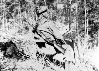 Charles Abel Corwin, artist painting scenery in the woods for the background mural of the Mule Deer Diorama Group. Rifle [gun] at his side. Captain Marshall Field Pacific Coast and Arizona Expedition Zoology July 1926. ID# CSZ52858