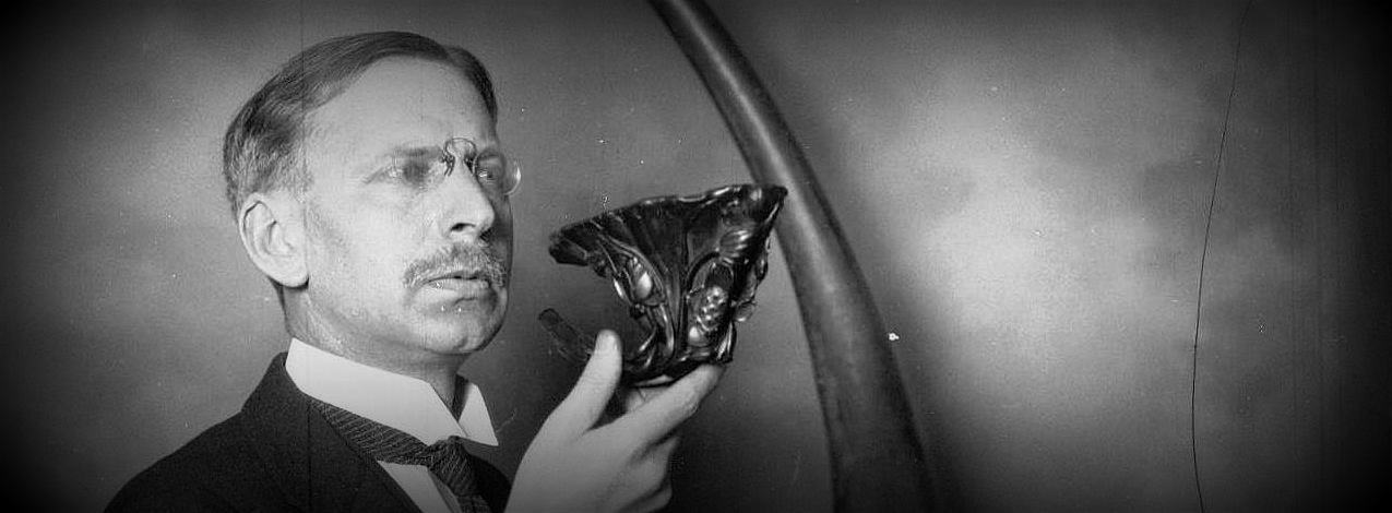 In a black and white photograph, a man wearing a pince-nez as well as a suit and tie holds up a carved horn.