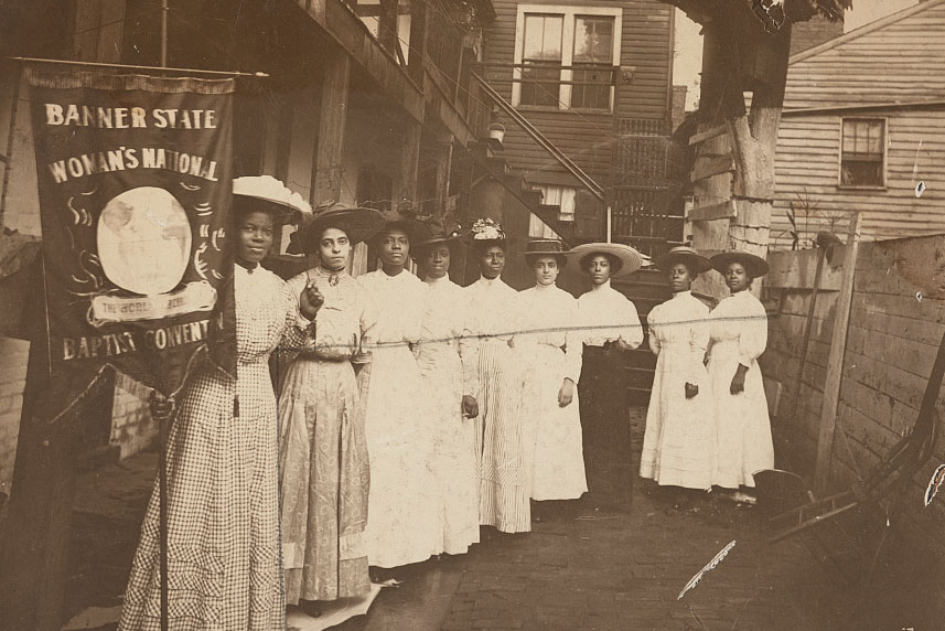 A group of African American women suffragists pose for a photo. On the left, closest to the camera, Nannie Helen Burroughs holds a banner that reads, Banner State Woman's National Baptist Convention.