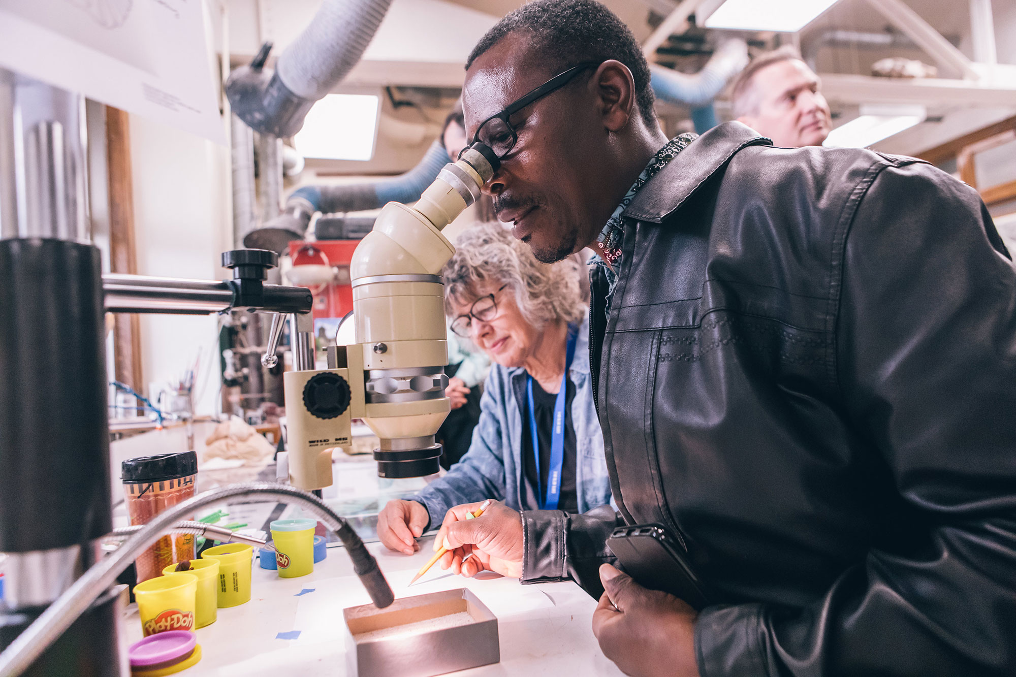 A man standing at a table peers into the eyepiece of a microscope to look at a specimen in a box. He holds a pencil in his right hand to sketch on a piece of paper on the table.