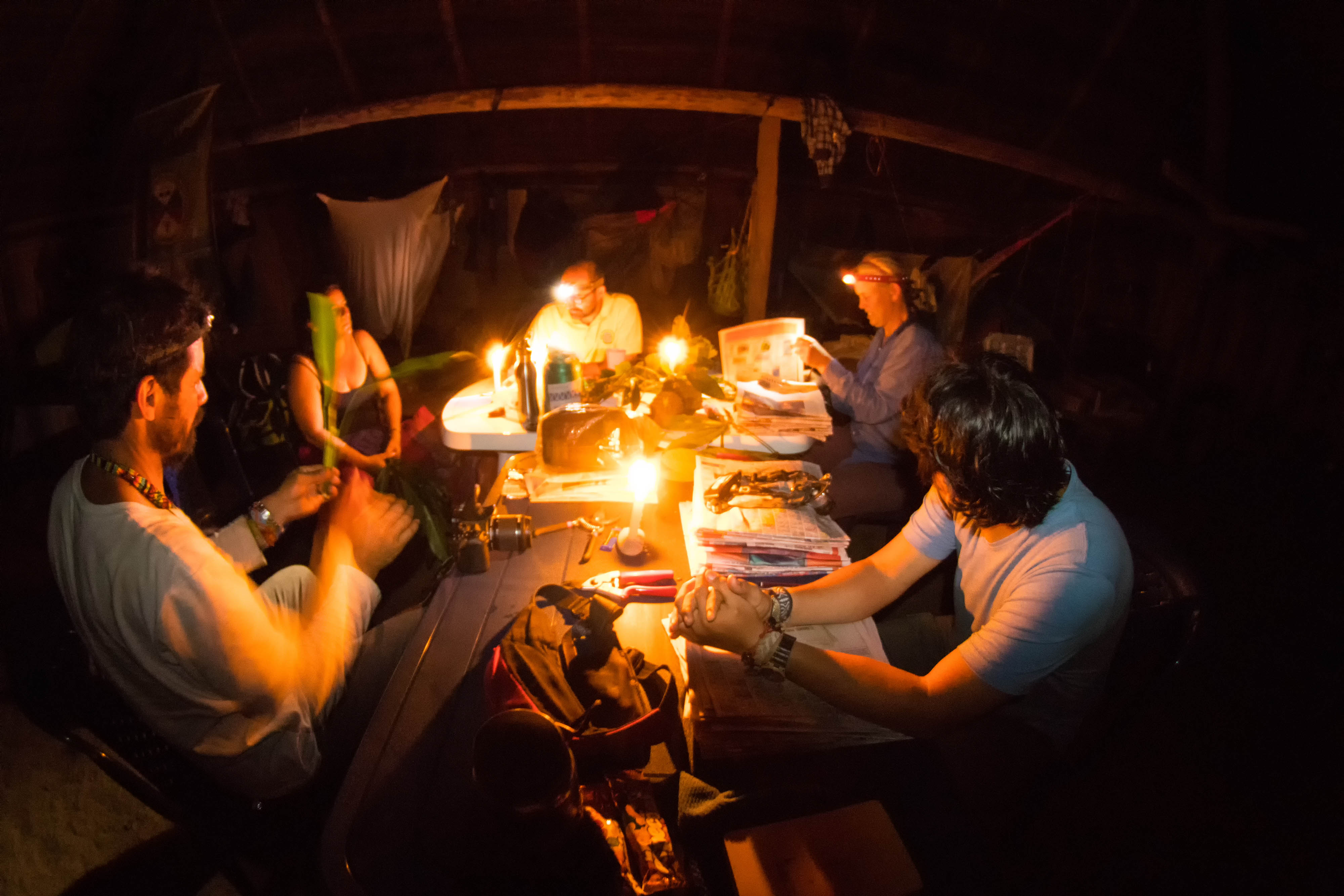 Five people sit around a table at night with several candles lit. Three of them also wear headlamps. They look through pieces of paper and notebooks, and one man holds up a plant specimen. Other items on the table include a camera, pliers, and a backpack.