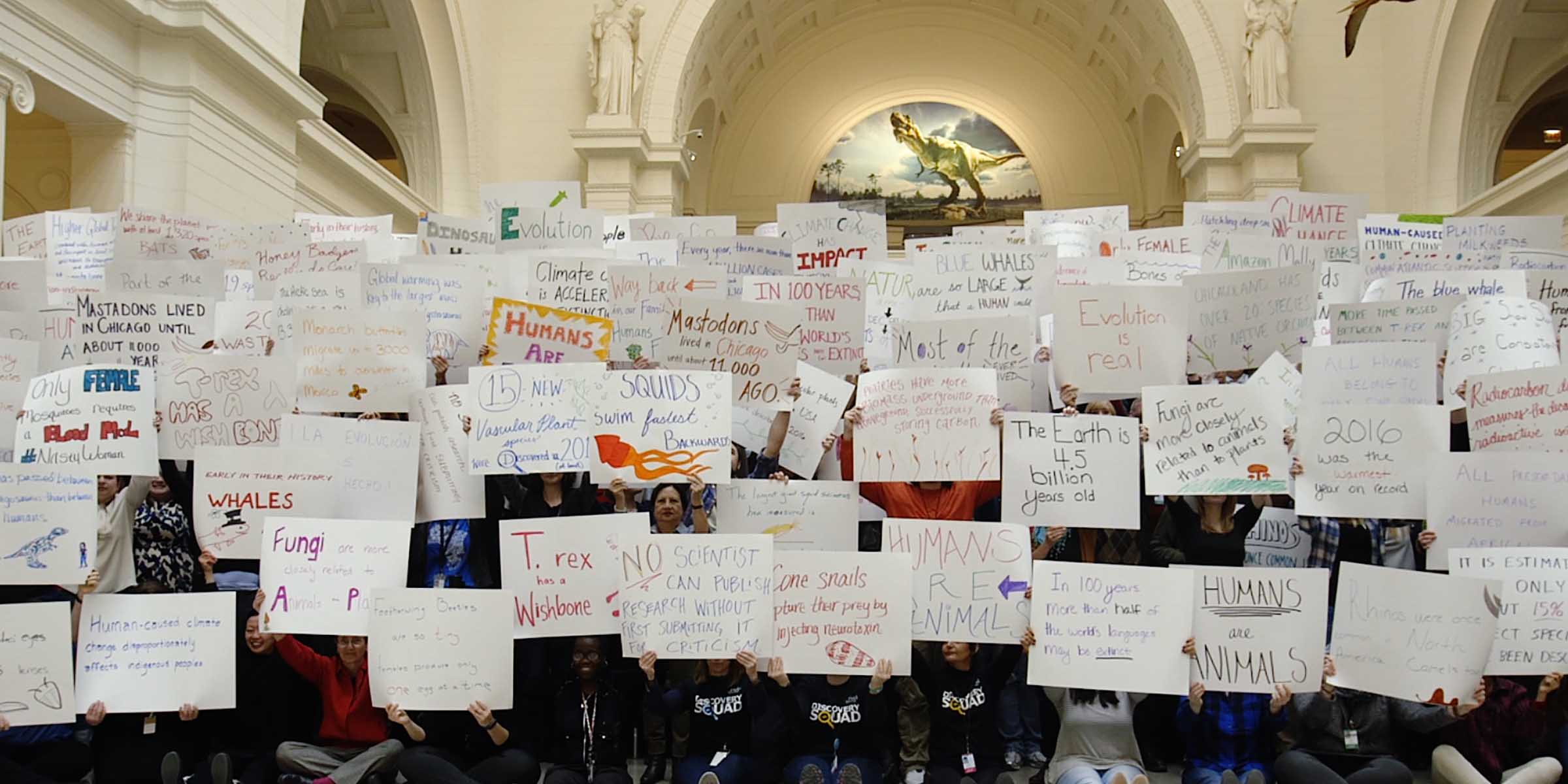 A group of about 150 people holding up signs with writing on them, inside a Neoclassical-style building