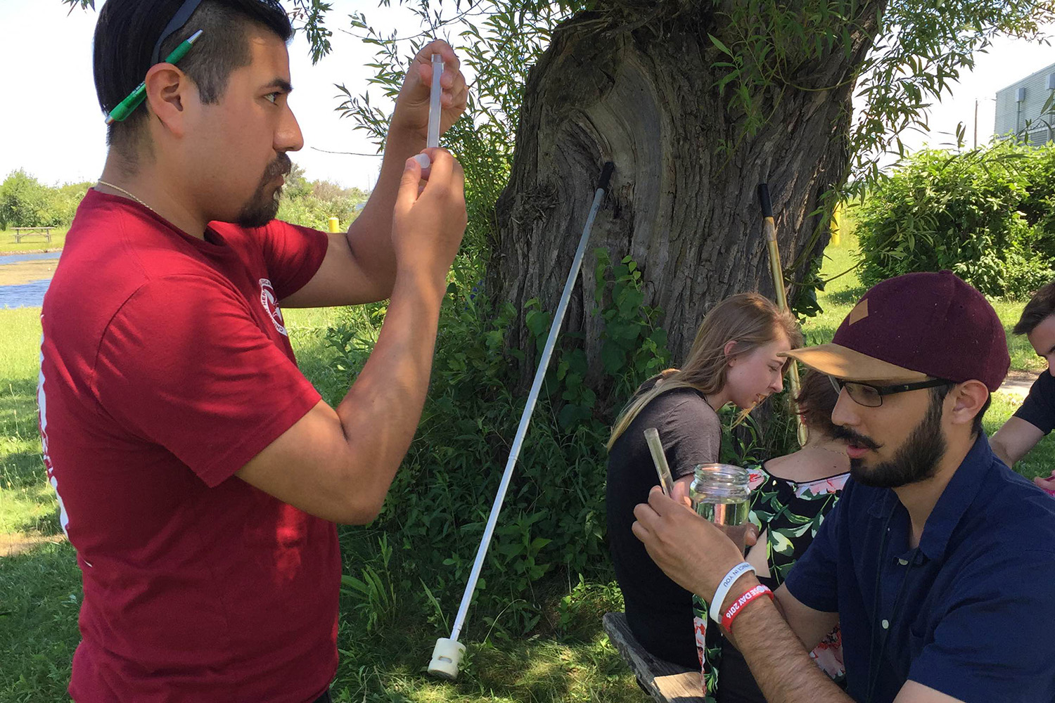 College-age students work together to document samples, one on the left side stands and others sit around a picnic table in an outdoor setting.