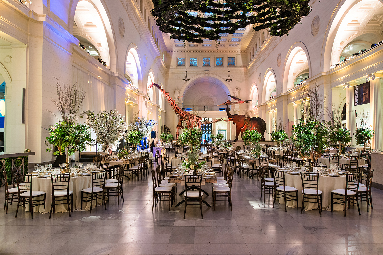 View of Stanley Field Hall during an event. Round and rectangular tables fill the floor in Stanley Field Hall. All tables are topped with large flower centerpieces and including seating and place settings for several guests.