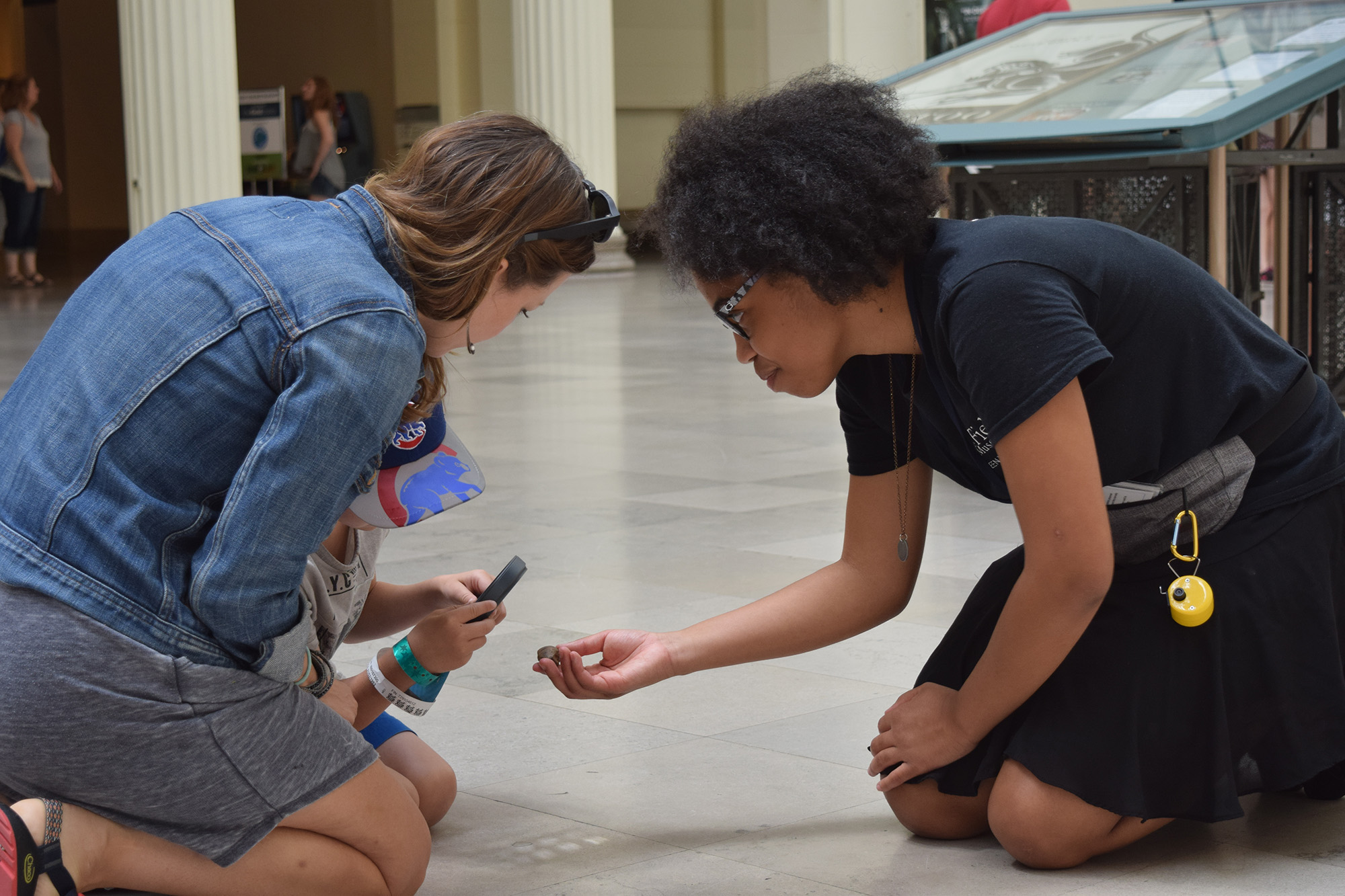Two teens kneel on a tile floor next to a child. They are all looking at a small object one of the teens is holding out in her hand.