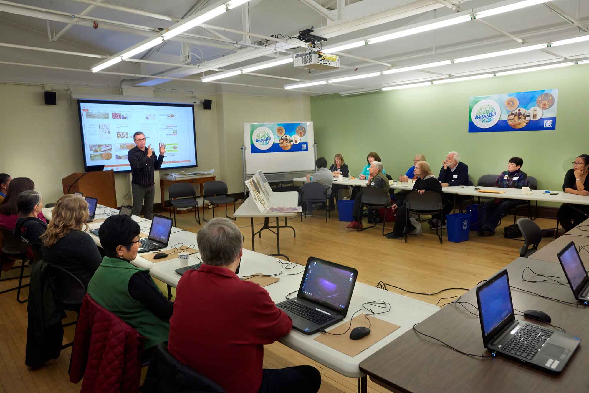 A group of volunteers gather in a classroom to digitize collections records. They sit at a set of tables arranged in a U shape. A scientists stands in front of the room, explaining the activities for the day.