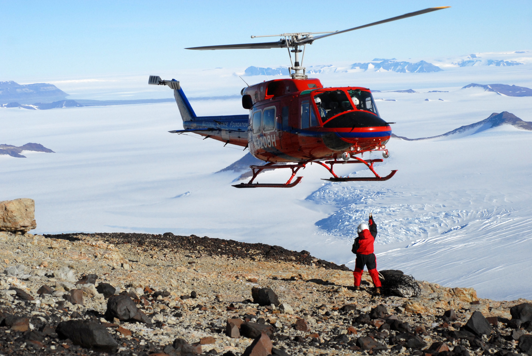 A red helicopter hovers close to the ground, which is rocky in the foreground and an expanse of snow in the background. A person dressed in a red snowsuit and helmet reaches a hand up towards the helicopter to attach a cargo net with fossils.