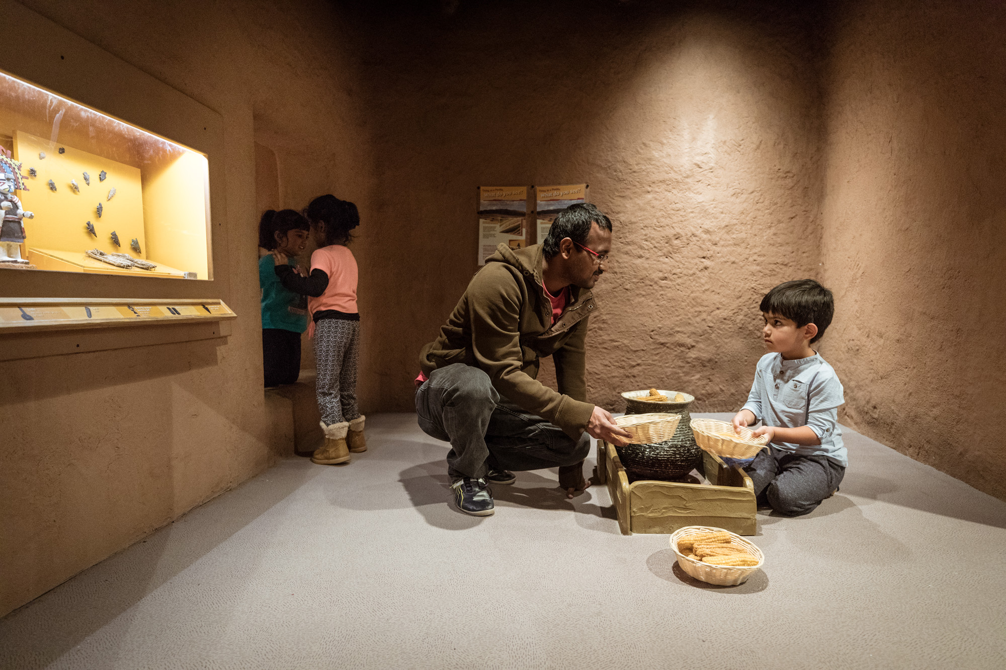 Inside a recreated pueblo in the Crown Family PlayLab, a child and an adult play with baskets holding fake corn. Two girls stand in the doorway nearby.