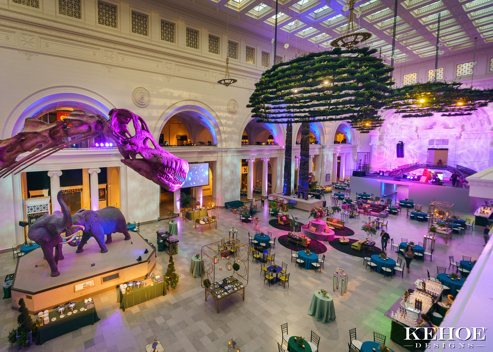 A view of a large museum hall arranged for a special event, seen from the second floor balcony. A dinosaur fossil head can be seen to the left, hanging gardens to the right.