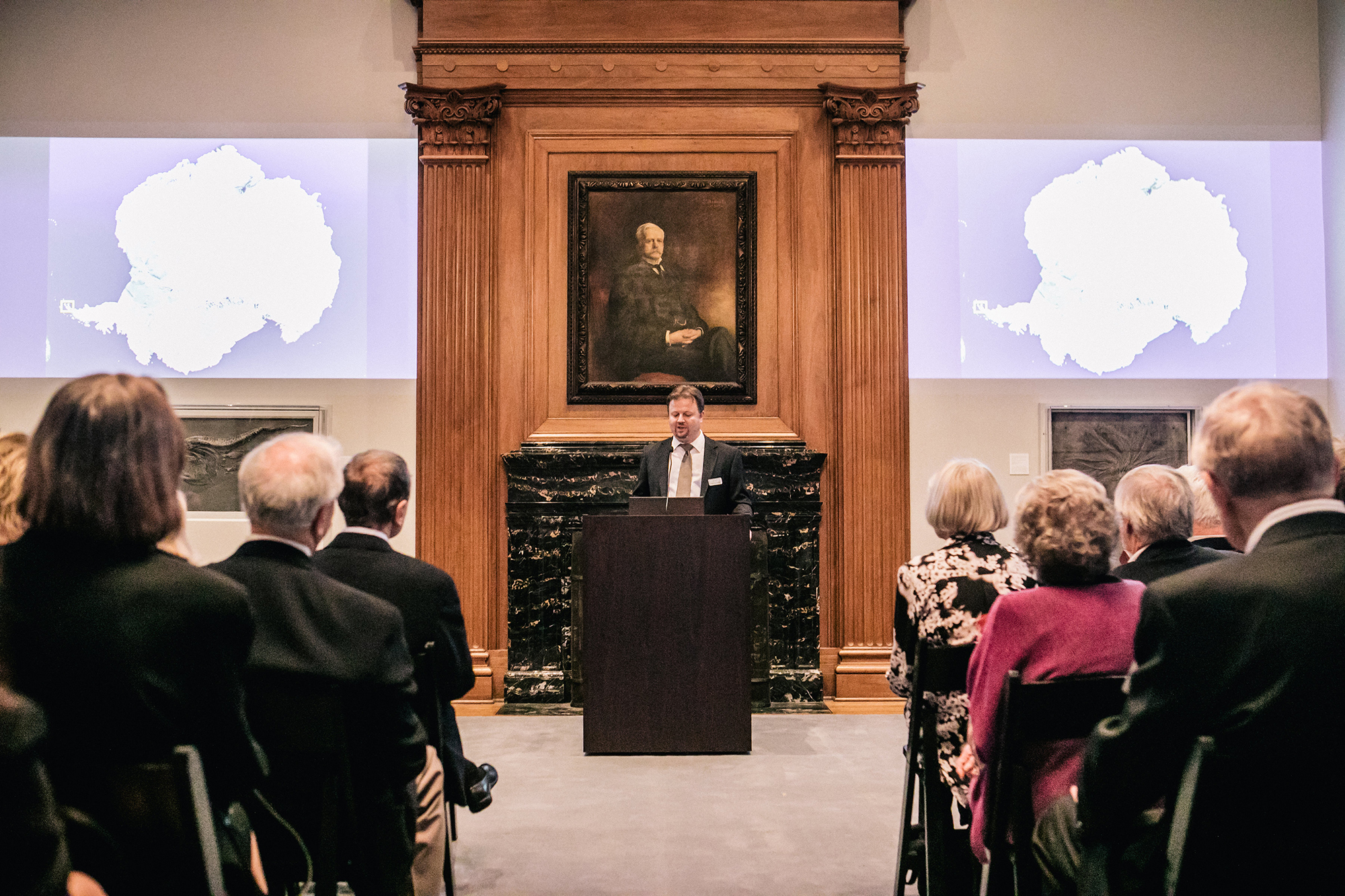 A man stands at a podium, which is placed in front of a black marble fireplace. The fireplace surround is a warm wood color, with a column on each side. Above the fireplace is a portrait of a man in academic robes. A presentation is projected the wall on either side of the fireplace, and men and women sit in rows of chairs watching the speaker.