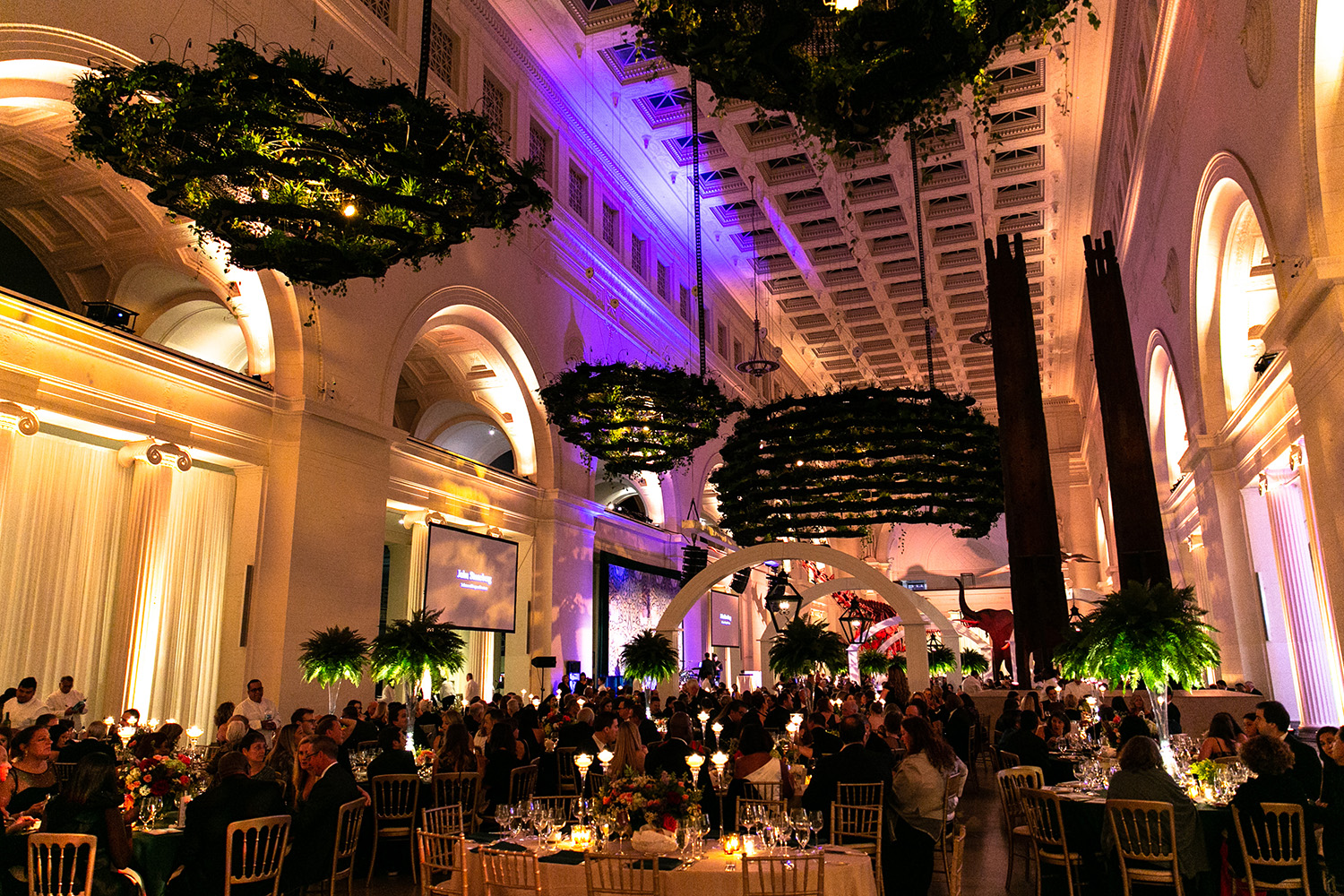 Stanley Field Hall set up for an evening gala event, with attendees seated at circular tables. Dramatic lighting highlights the hanging gardens, and screens displaying a presentation are visible towards the back of the frame.